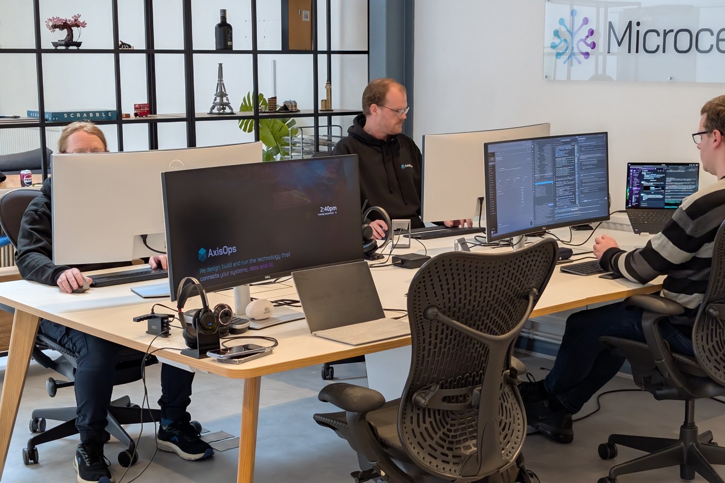 Three engineers at a shared desk in the AxisOps office, screens up, the Microcelium sign on the wall behind them.